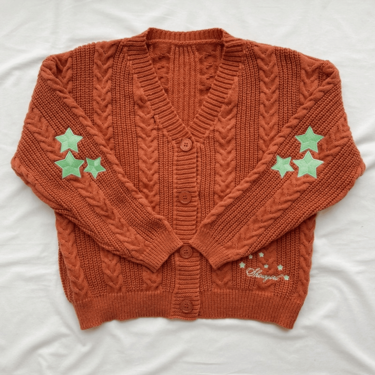 An orange cable-knit cardigan sweater with three light green star patches on each sleeve and a small embroidered script on the lower right front, laid flat on a white fabric background.
