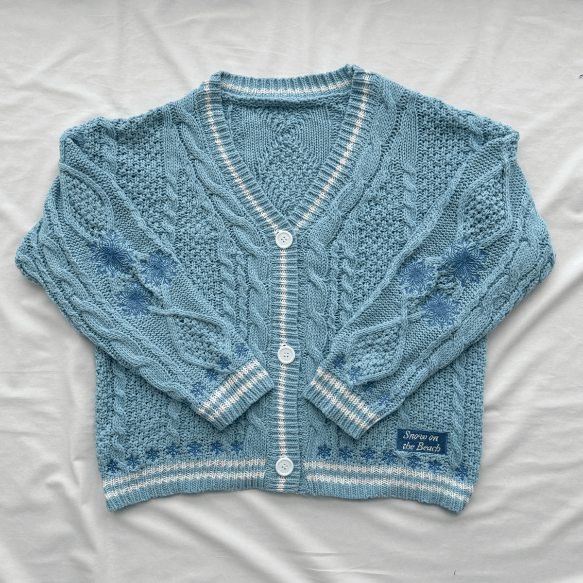 A light blue cable-knit cardigan sweater with white striped trim and white buttons, laid flat on a white fabric background. A small woven label on the lower right reads 'Snow On The Beach'.