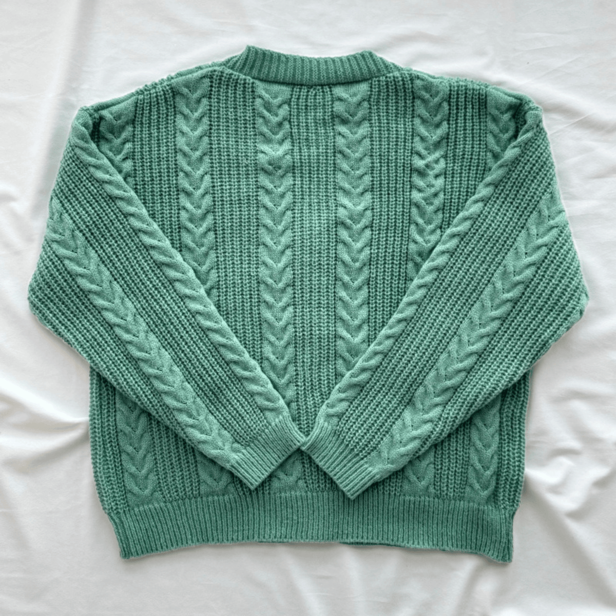 A sage green cable-knit sweater with a crew neck and long sleeves, laid flat on a white fabric background.