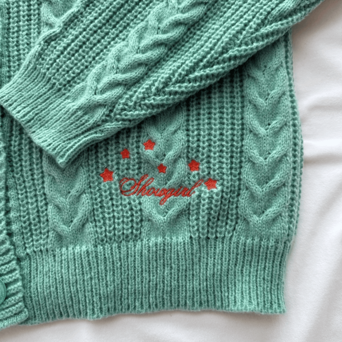 A mint green cable-knit cardigan sweater with red embroidered stars and the word 'Showgirl', laid flat on a white fabric background.