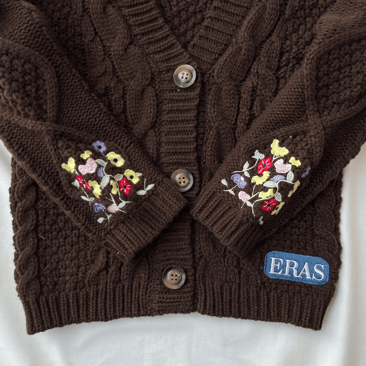 Brown cable knit cardigan with floral embroidery on sleeves and 'ERAS' logo on a white background