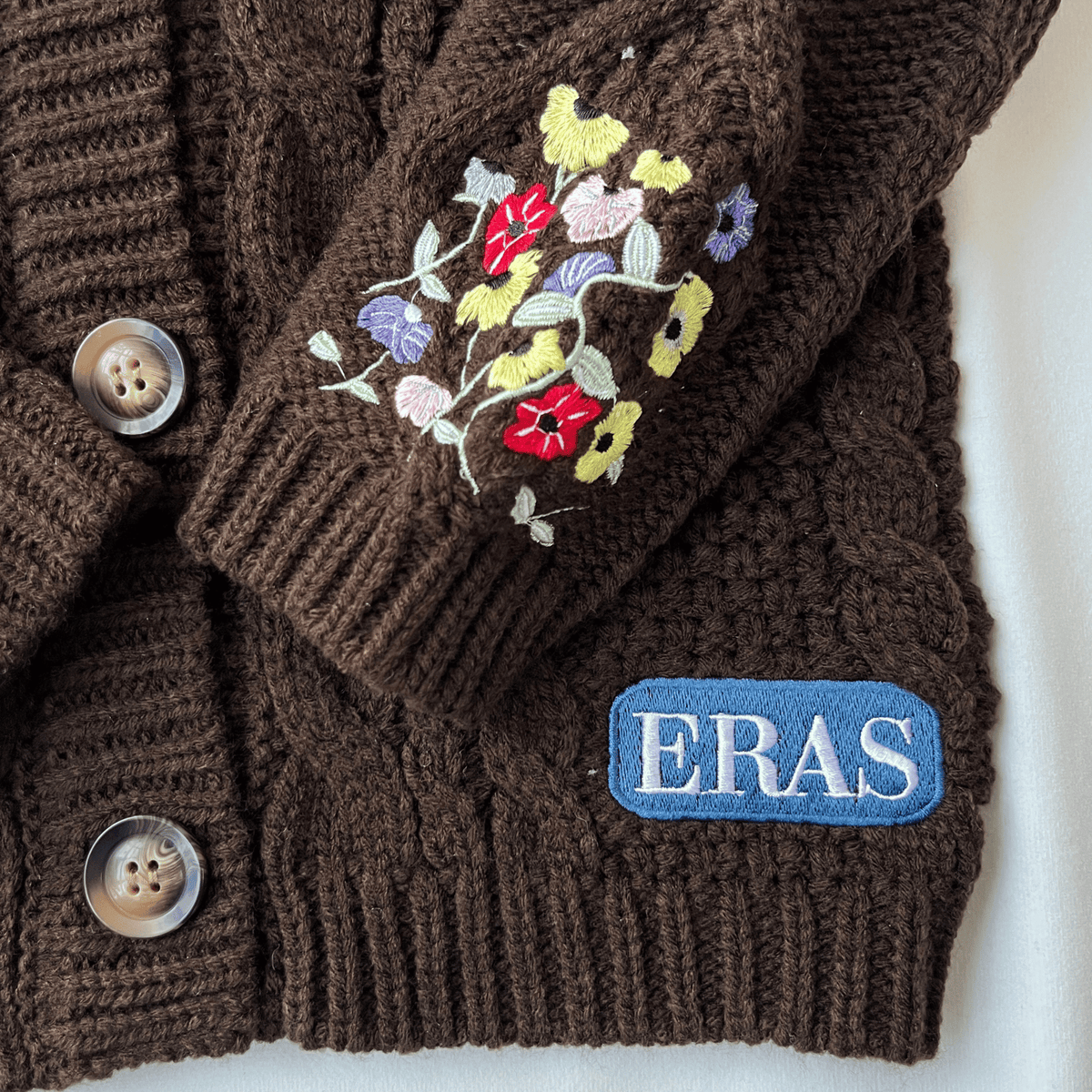 Brown knitted sweater with floral embroidery and 'ERAS' logo on a white background