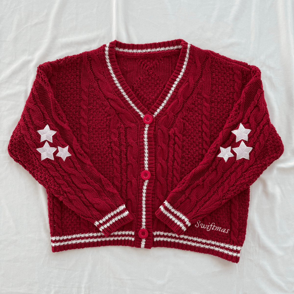 A red cable-knit cardigan sweater with white trim, featuring three white stars on each sleeve and a white embroidered 'Swiftmas' brand name on the lower right front. The cardigan is laid flat on a white fabric background.